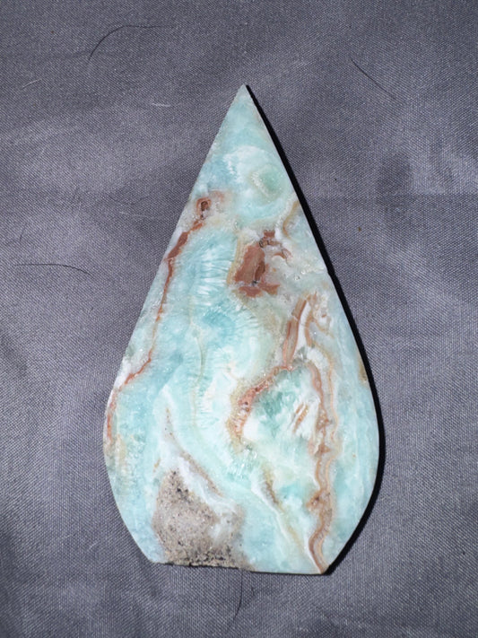 A polished teardrop-shaped stone slab with natural marbling in light blue, white, and brown, displayed on a textured dark gray fabric background.