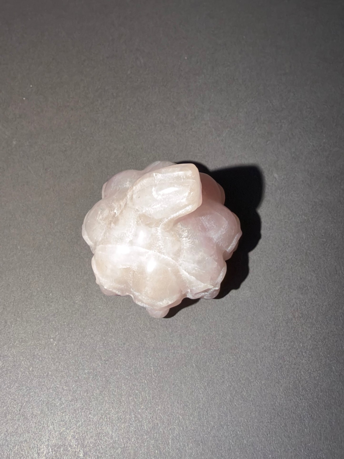 A pale pink carved stone or crystal cluster with a rough, organic shape, photographed on a smooth dark gray surface with a cast shadow.