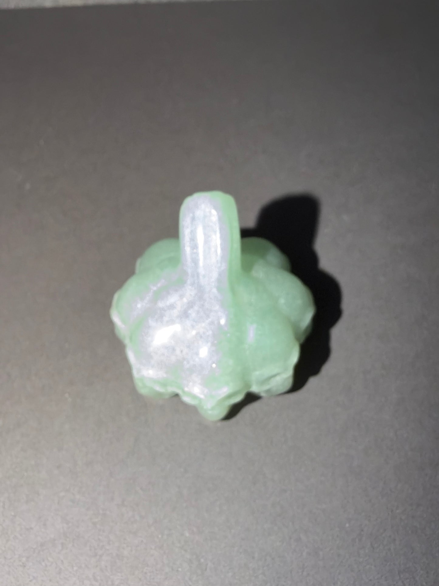 A small carved pale green jade pendant with a loop at the top, photographed on a dark gray surface with a soft shadow.