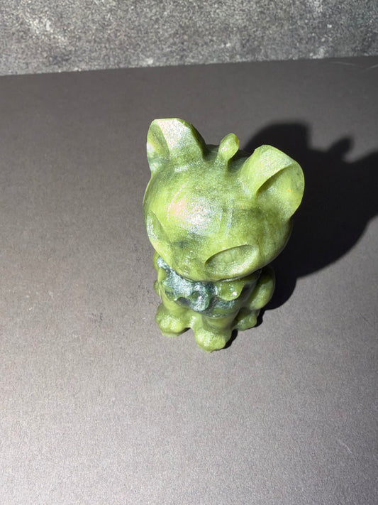 A small green stone figurine of a cartoon cat or kitten, photographed on a dark gray surface with a cast shadow.
