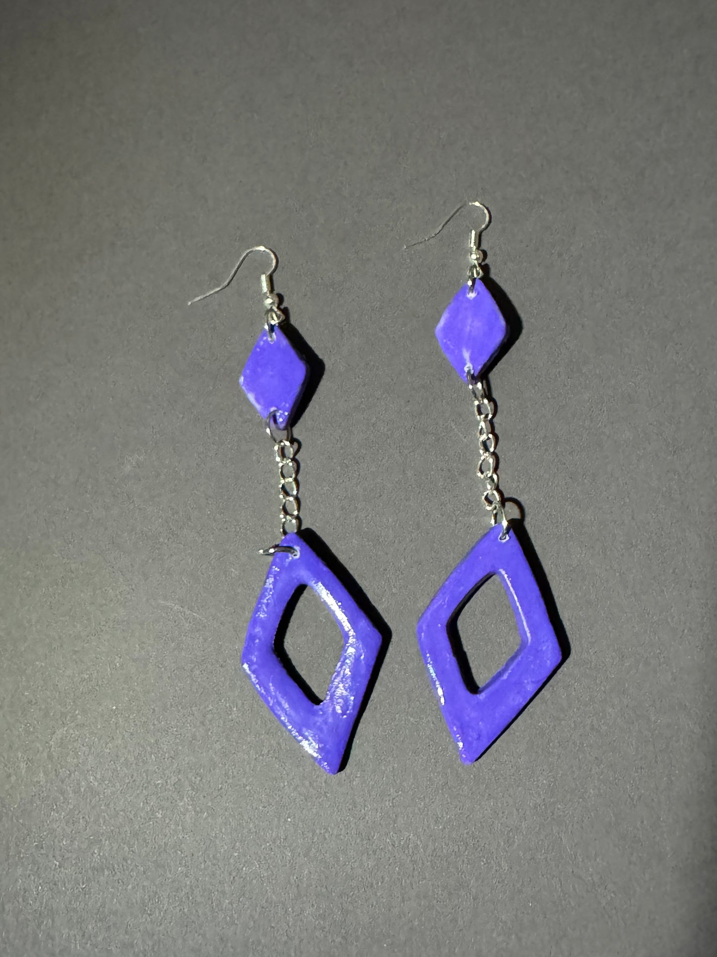 A pair of handmade dangle earrings featuring textured, glossy purple geometric shapes and silver-tone metal hooks, displayed on a neutral gray background.