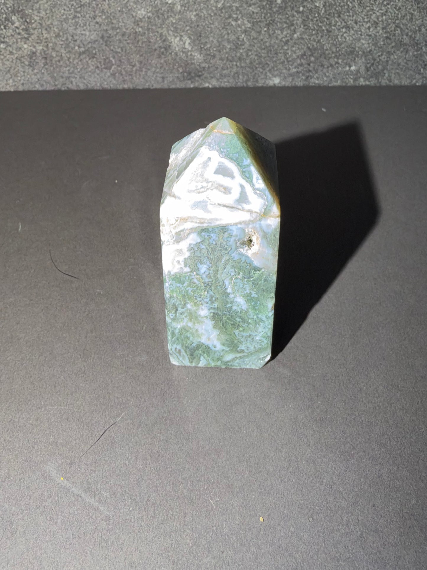 A polished natural crystal point, likely jadeite, featuring marbled green, white, and light gray patterns, standing upright on a dark gray surface.
