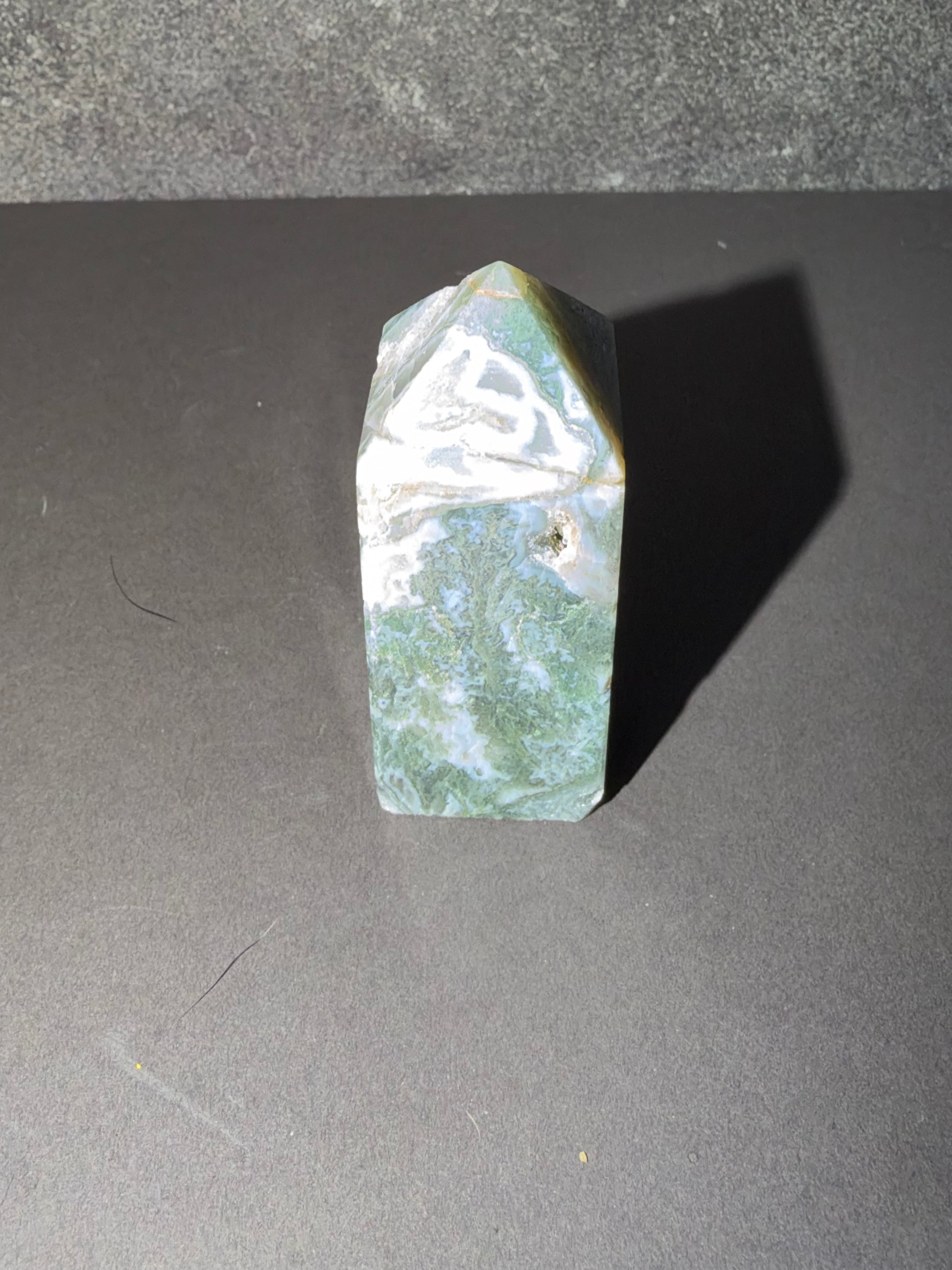 A polished natural crystal point, likely jadeite, featuring marbled green, white, and light gray patterns, standing upright on a dark gray surface.