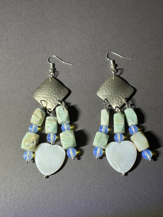 A pair of handmade dangle earrings featuring hammered silver-tone metal, light green marbled beads, blue and white striped beads, and white mother-of-pearl heart pendants, displayed on a dark gray background.