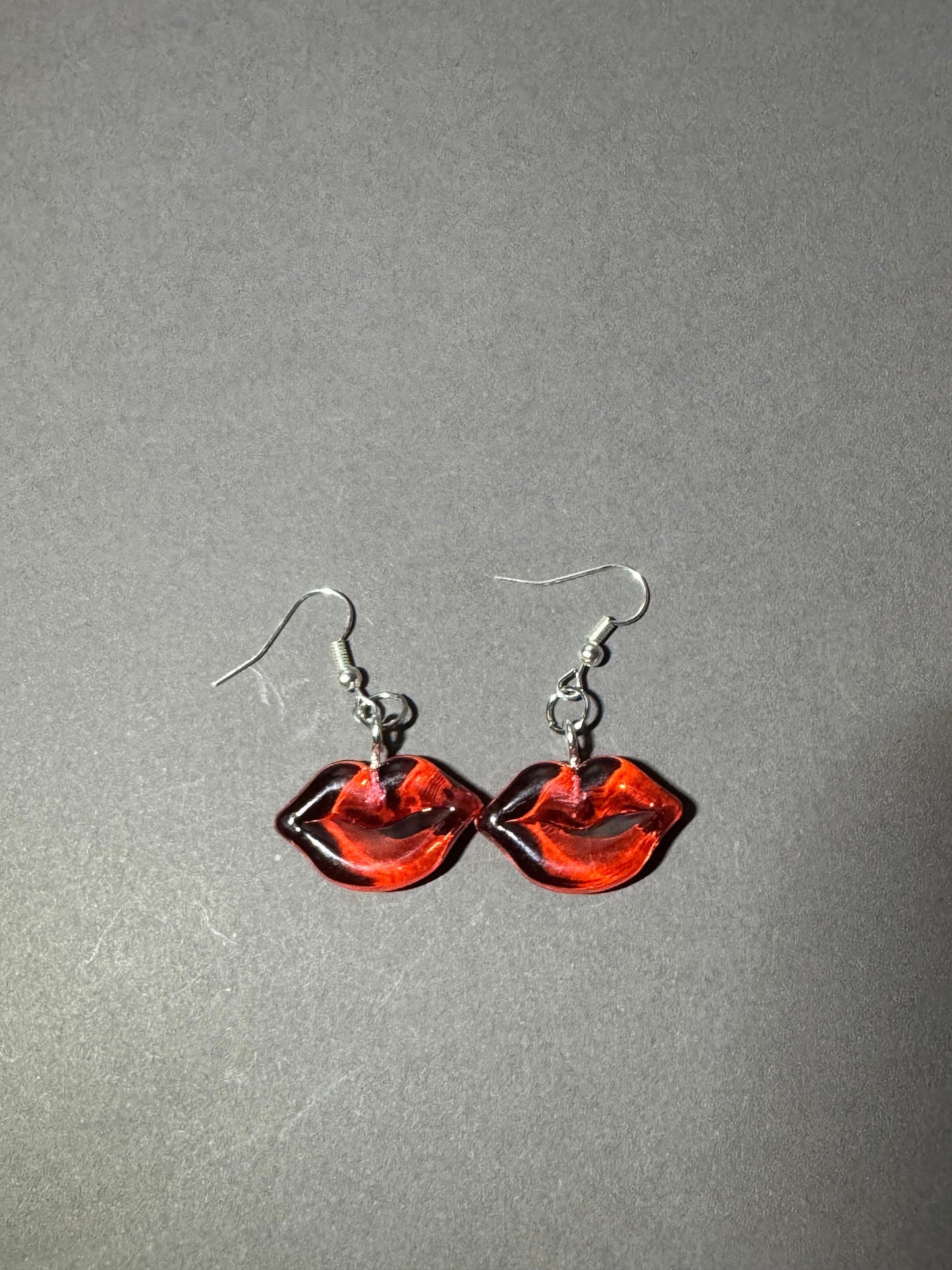 A pair of red lips-shaped dangle earrings with silver fishhook wires, displayed on a textured gray background.