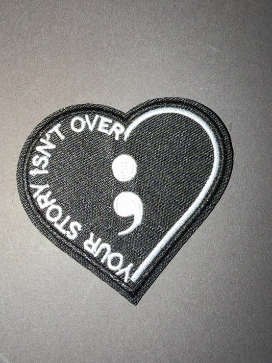 A heart-shaped embroidered patch with a black background, white semicolon symbol, and the text 'YOUR STORY ISN'T OVER!' stitched along the left side, shown on a plain gray surface.