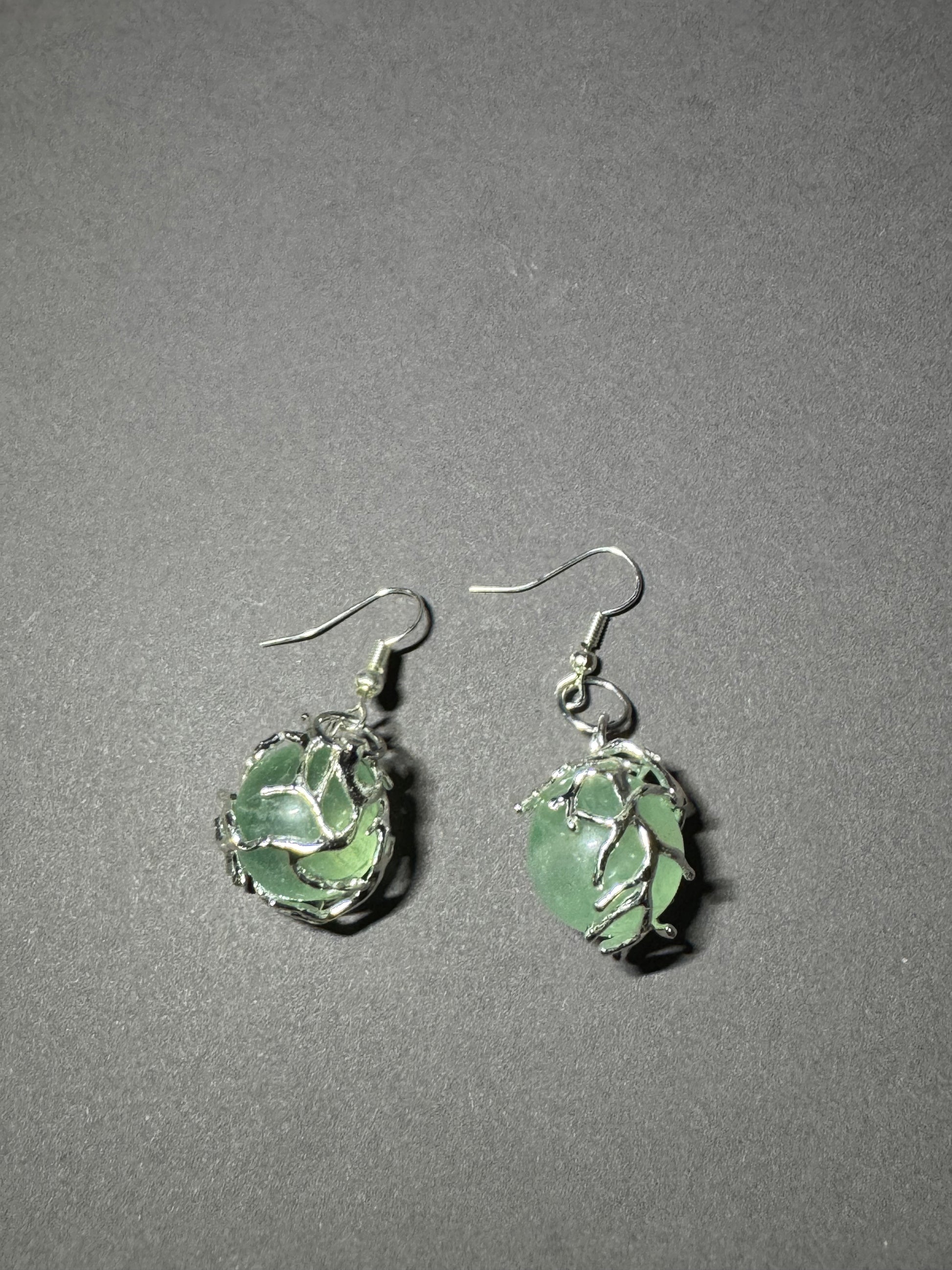 A pair of handmade dangle earrings featuring green sea glass wrapped in silver wire, suspended from silver fishhook wires, displayed on a textured gray background.