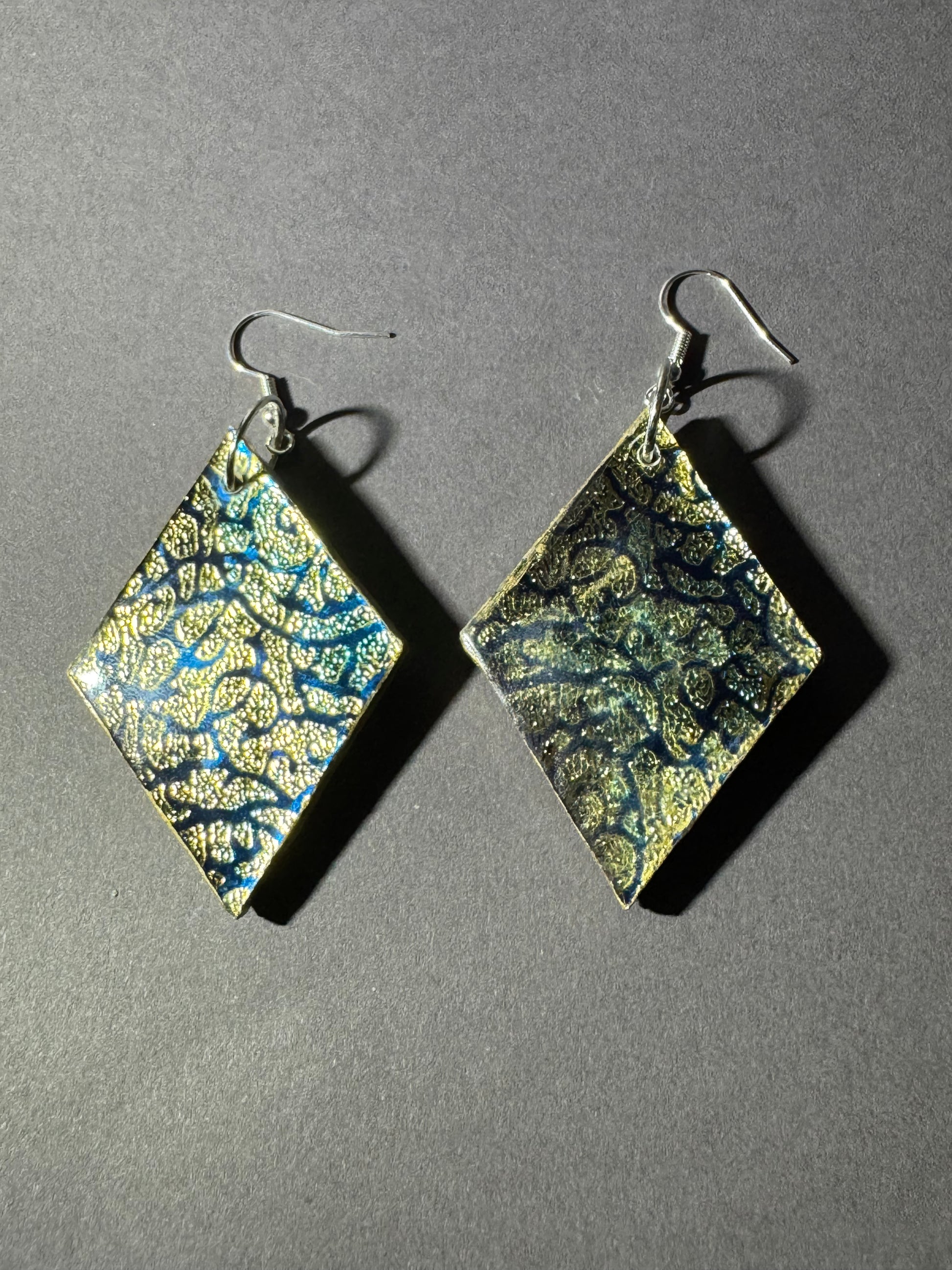 A pair of diamond-shaped dangling earrings with a marbled pattern of gold, black, and blue, suspended from silver fishhook wires on a dark gray background.