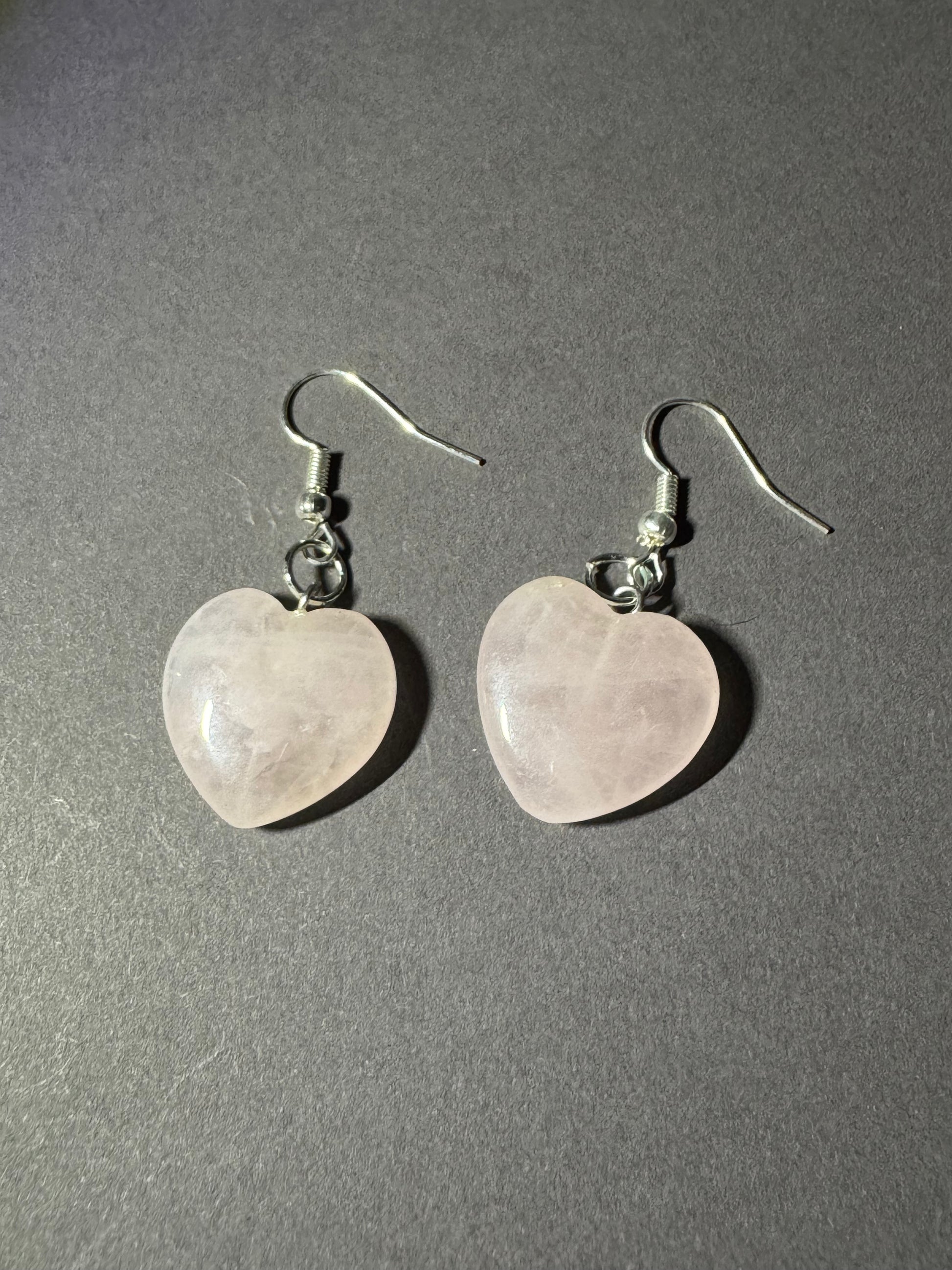 A pair of heart-shaped rose quartz drop earrings with silver fishhook wires, displayed on a textured dark gray surface.