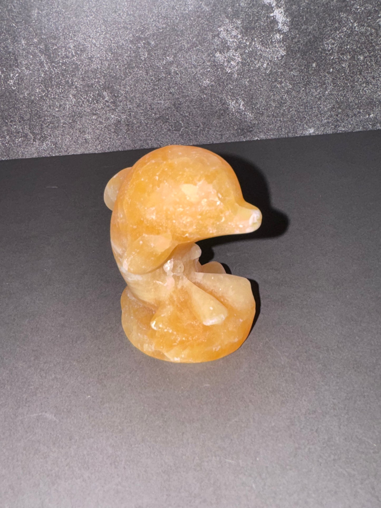 A small carved figurine of a dolphin or whale head and upper body made from translucent orange-yellow stone, standing on a dark gray surface against a textured dark gray background.