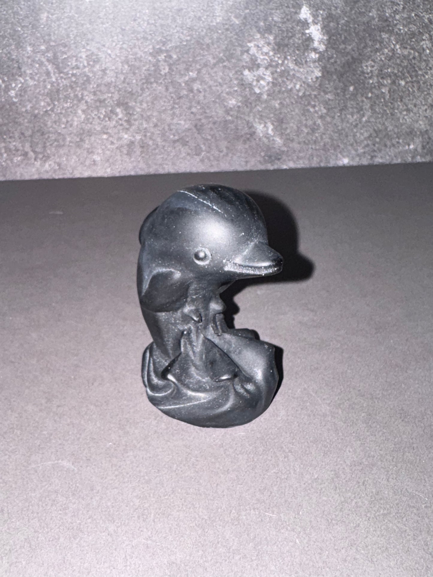 A small black dolphin figurine with a textured, matte finish, standing on a gray surface against a gray background.
