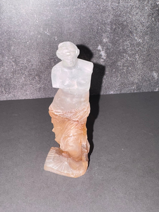 A small translucent figurine of a classical female bust and draped figure, made from layered milky white material on top of warm peachy-brown material, standing on a dark gray surface against a textured dark gray backdrop.