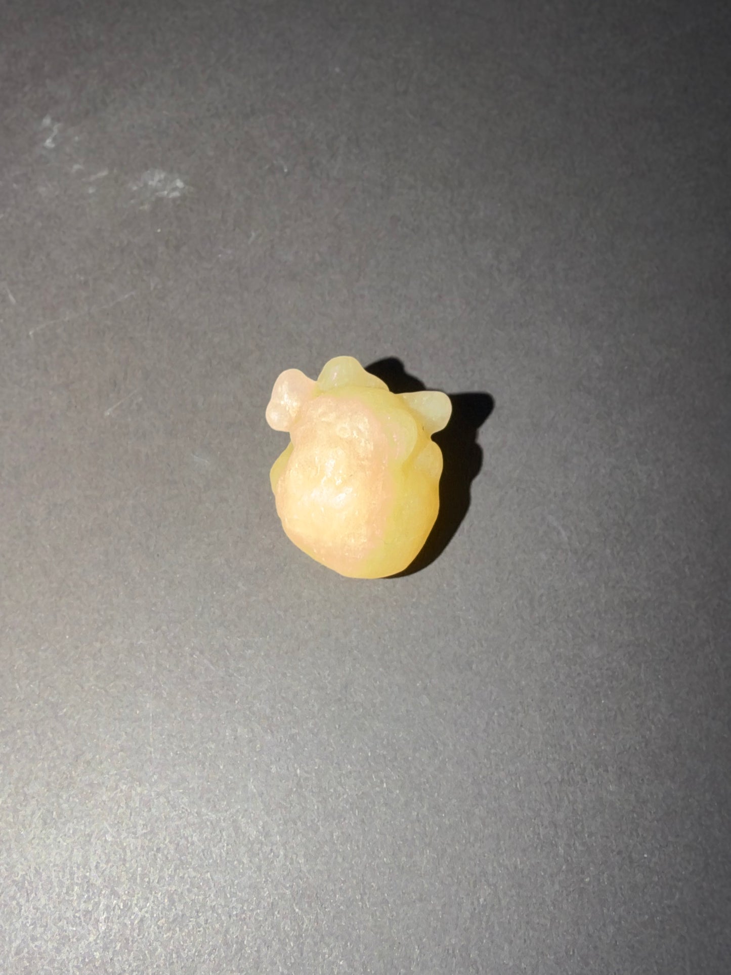 A small pale yellow translucent anatomical heart stone, placed on a dark gray surface.