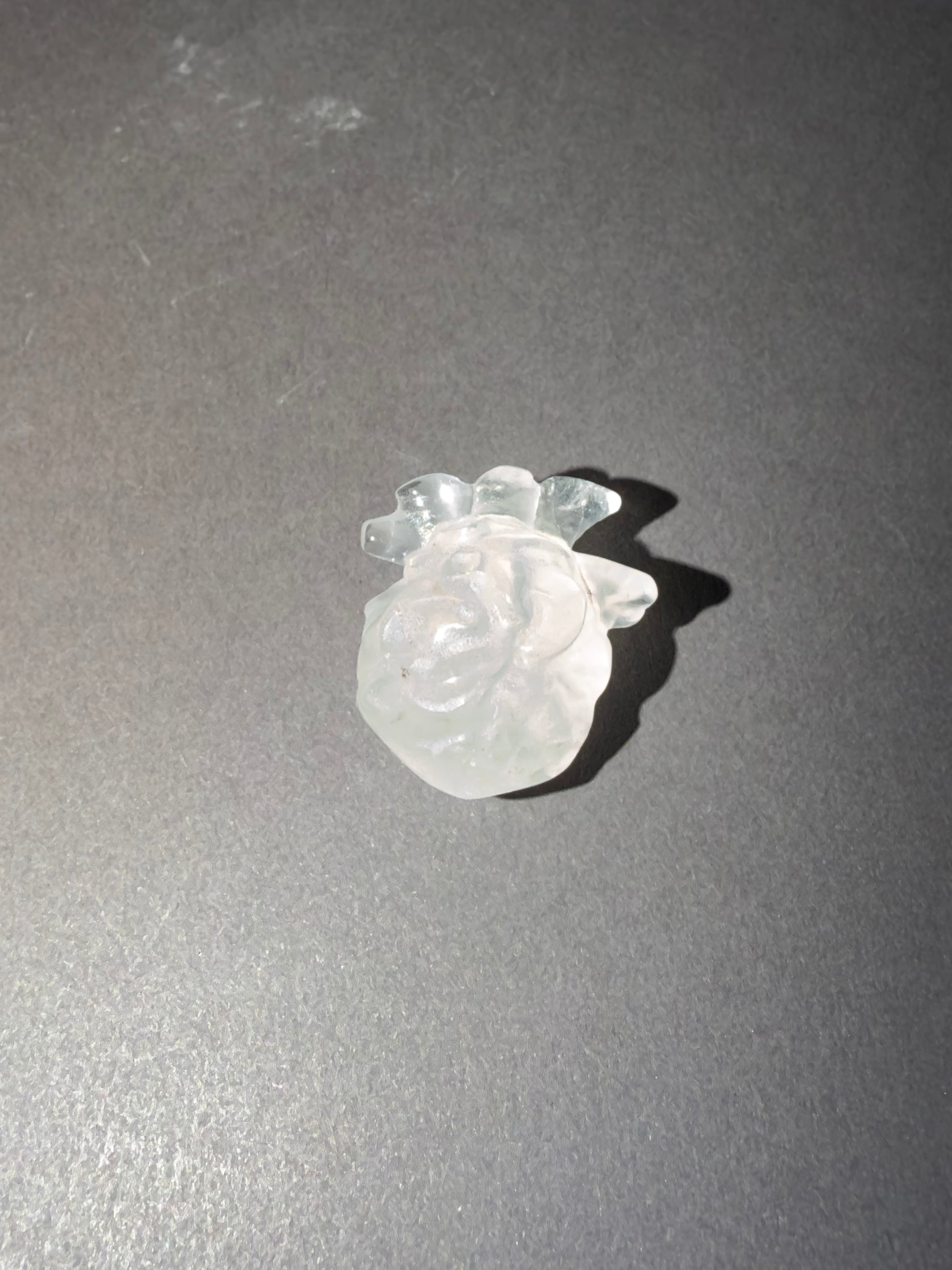 A small carved translucent white stone figurine, of an anatomical heart, placed on a dark gray surface with a cast shadow.