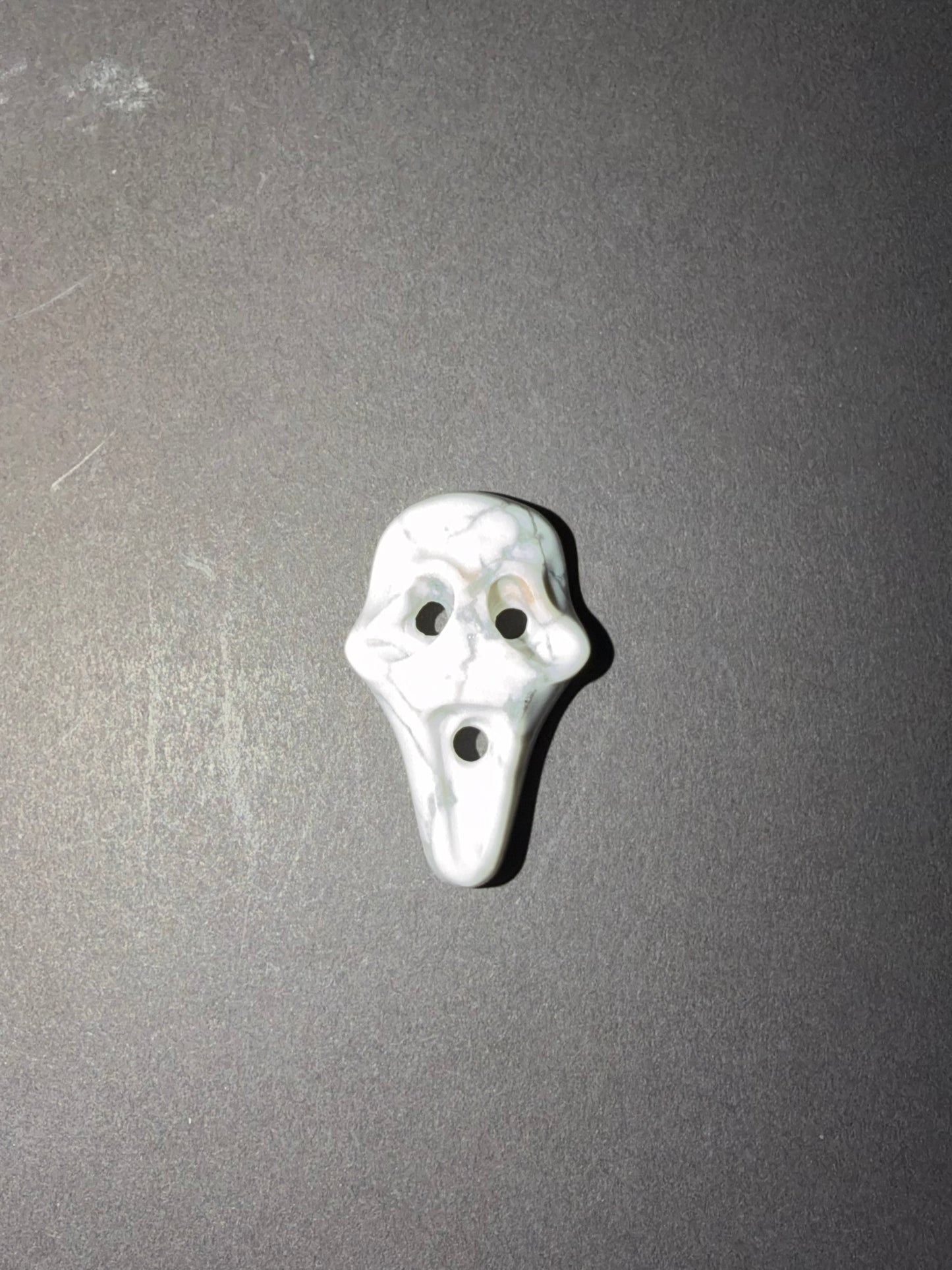 A small white and gray stone shaped like a ghost face with three holes, shown on a dark gray textured background.