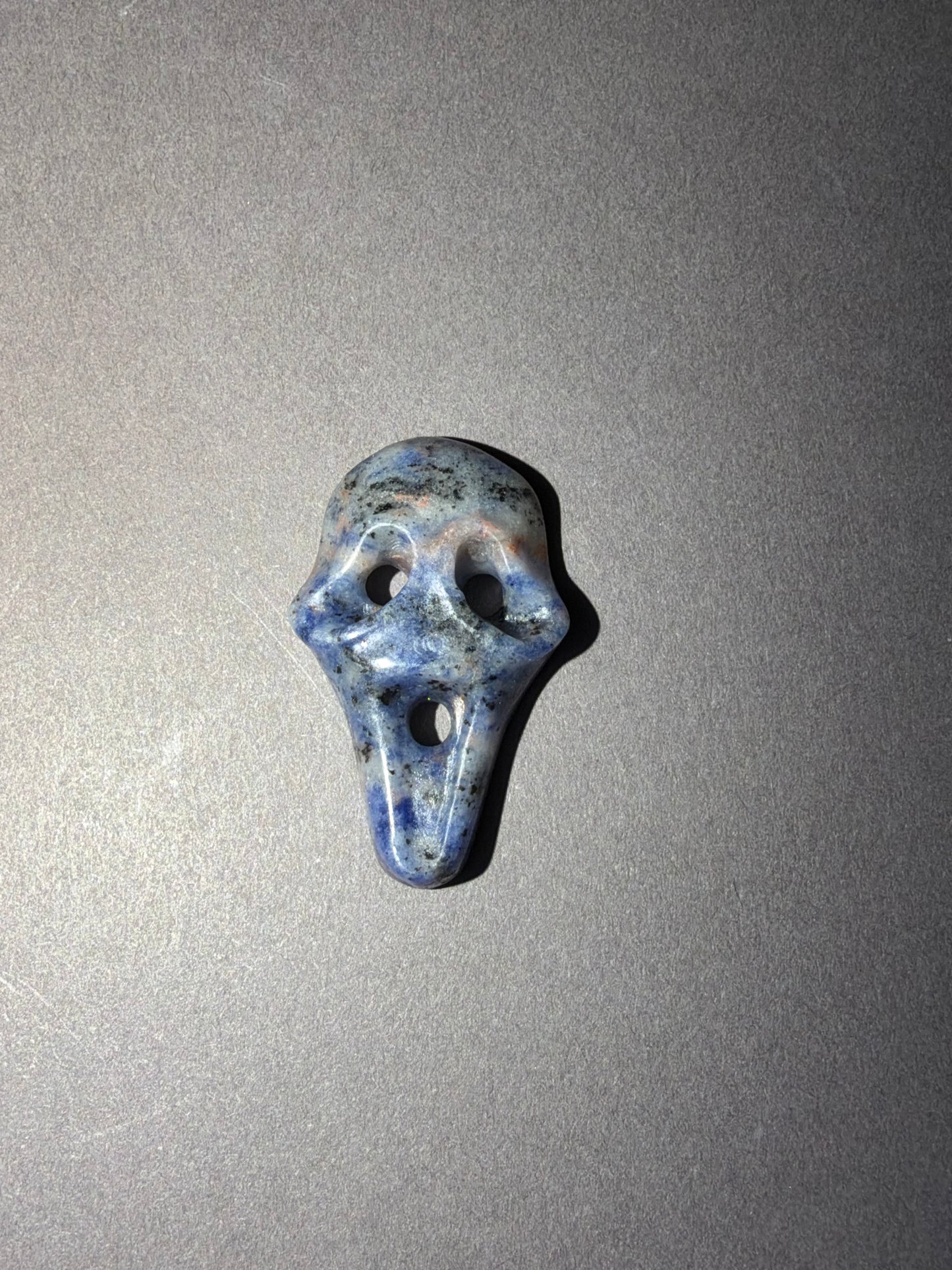 A small carved stone shaped like a ghost face, featuring blue, gray, and white marbled patterns, set against a plain gray background.