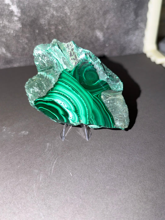 A raw green malachite crystal specimen with striking concentric banding, displayed on a clear acrylic stand against a dark gray background.