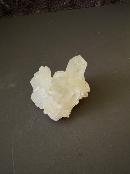 A raw white crystal cluster with multiple translucent points, resting on a dark gray surface.