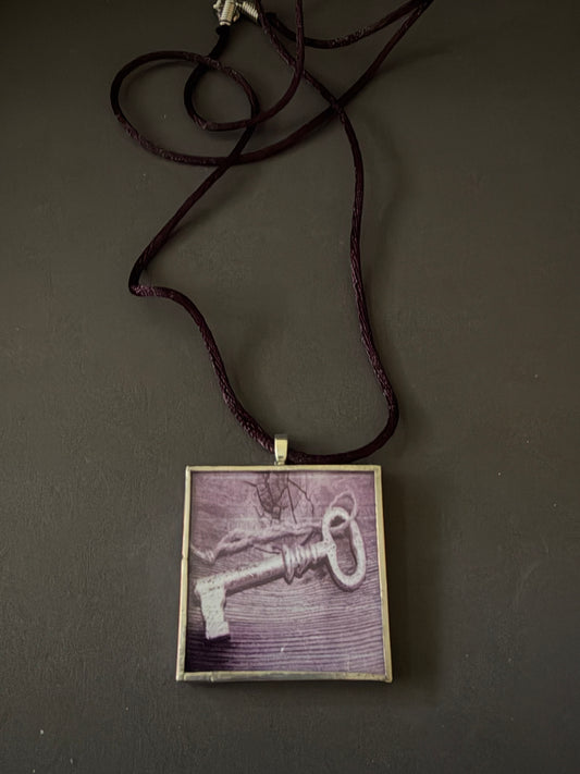 A square pendant necklace featuring a vintage-style key illustration in muted purple and white tones, suspended from a dark plum cord on a dark gray background.