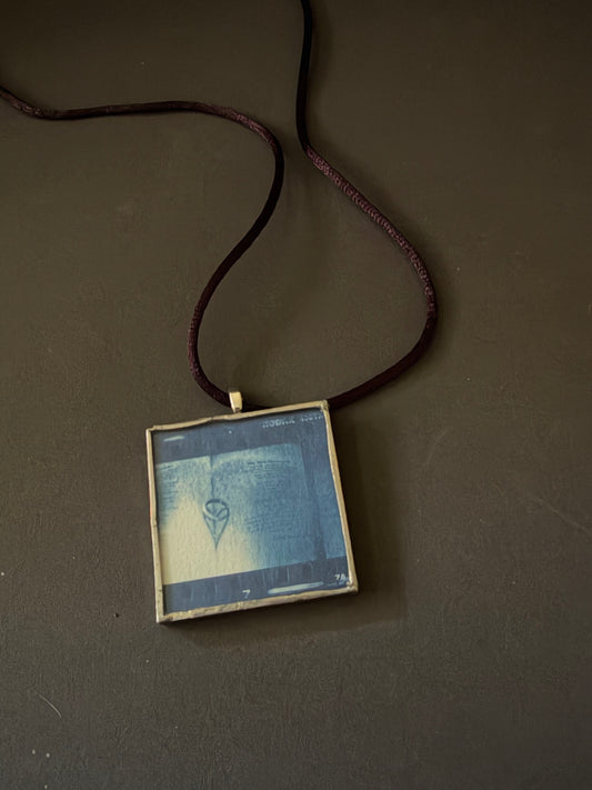 A square pendant necklace featuring a blue-toned graphic print in a glass  panel, suspended from a dark burgundy cord against a dark brown background.