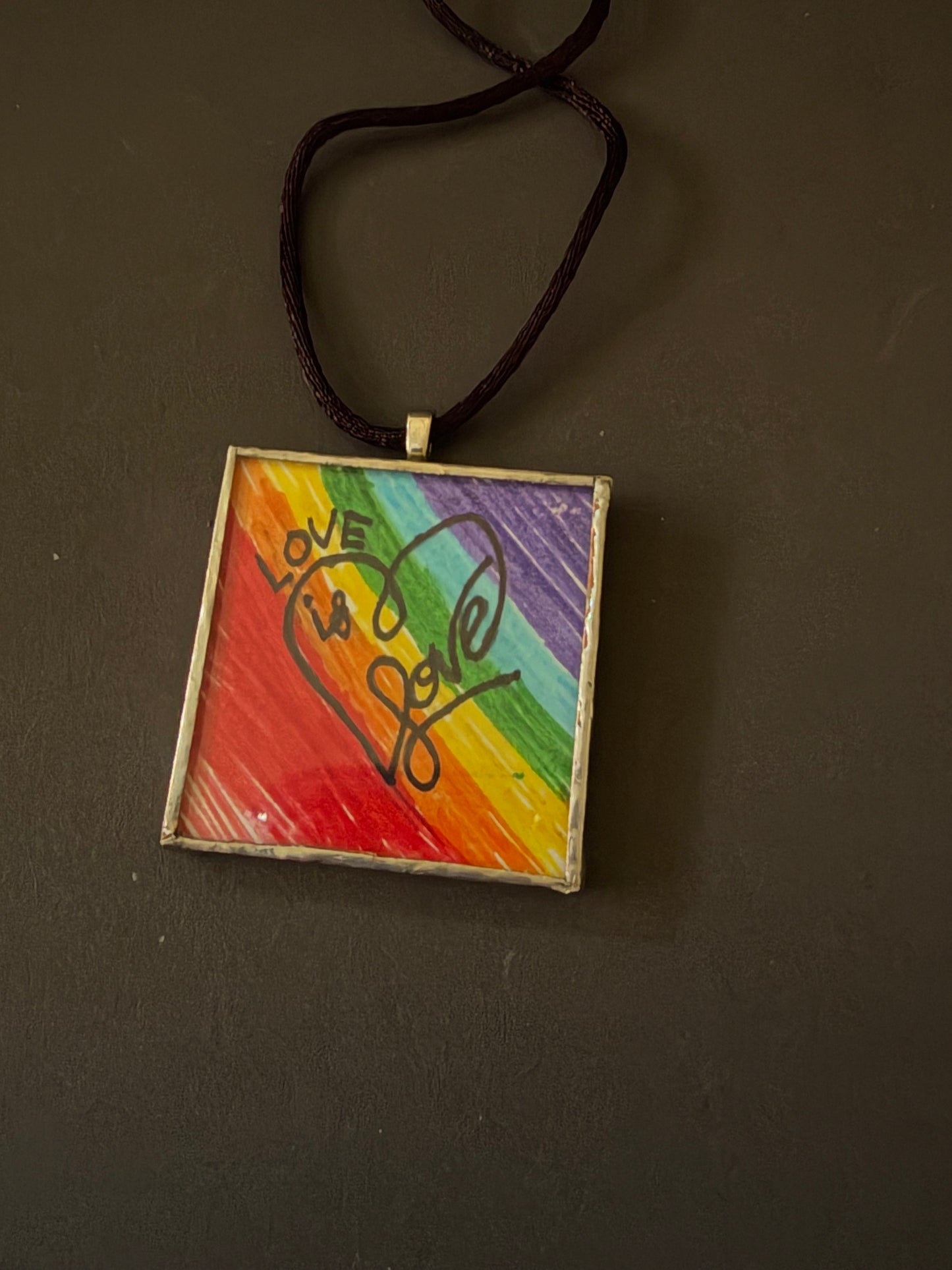 A square pendant necklace featuring a colorful rainbow background with a heart design and the words 'LOVE is love' in black script, suspended from a dark brown cord on a dark gray background.