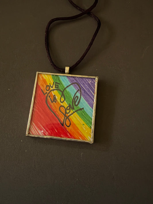A square pendant necklace featuring a colorful rainbow background with a heart design and the words 'LOVE is love' in black script, suspended from a dark brown cord on a dark gray background.